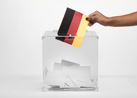 Eine Person wirft einen Zettel in den Farben der deutschen Flagge in eine durchsichtige Wahlurne